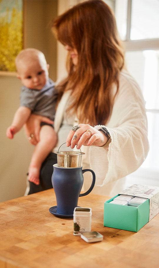 a woman holding a baby in one arm takes an infuser out of a tea cup