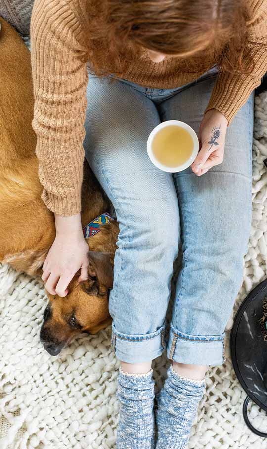 a girl holds a tea cup and pets a dog on a bed