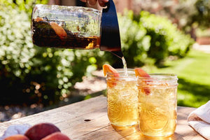 a pitcher pours iced tea into two glasses with peaches and iced tea