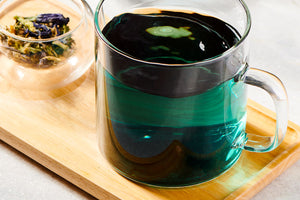 a clear mug with blue tea in it sits on a board with a dish holding butterfly pea flower tea