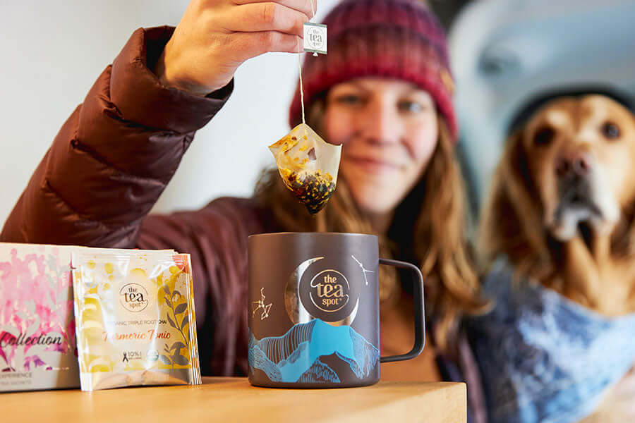 a woman holds a tea bag over a tea mug with a dog sitting beside her 