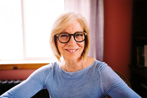a woman with blonde hair and glasses smiles in front of a window