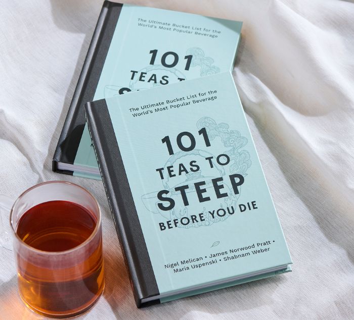 two books titled '101 Teas to Steep Before You Die' sit next to a brewed cup of tea on a white cloth surface