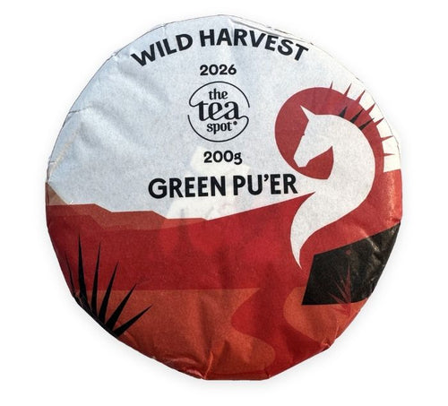 A single wrapped tea puer cake on a plain white background. The circular wrapper is white at the top and red at the bottom, with black text reading 'WILD HARVEST 2026 the tea spot 2005 GREEN PU'ER' and a stylized white horse head on a red shape.