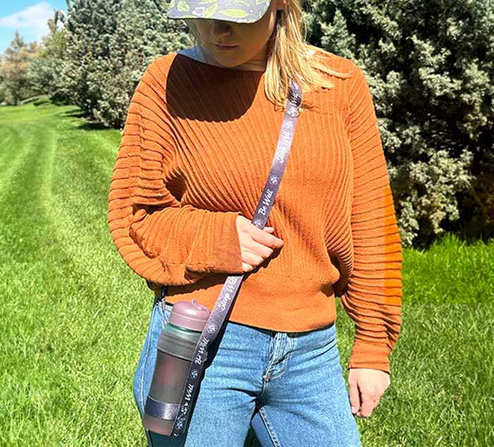 a person stands in a field with a water bottle strap across their body holding a water bottle