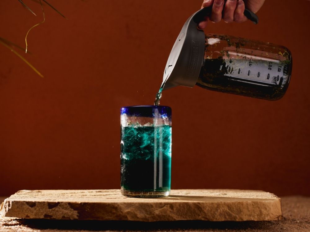 Person pouring blue tea from a measuring cup with a craft cold brew filter into a glass on a wooden surface with a brown background.