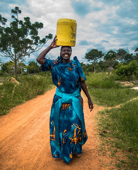 a woman walks on a path holding a yellow container of clean water on her head