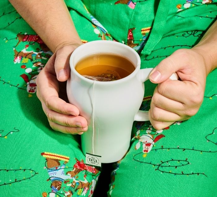 a person holds a tea mug in their lap with a tea bag steeping in it with a tag that reads the tea spot