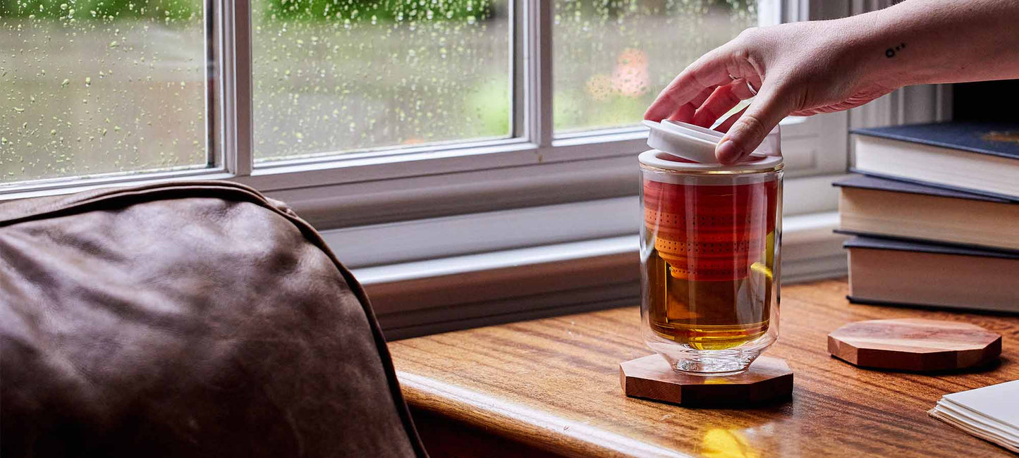 a hand pulls a tea steeper out of a glass tea cup sitting on a shelf near a window
