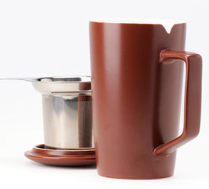 a brown ceramic tea mug sits in the foreground with a stainless steel tea infuser and lid behind it