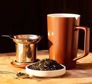 a brown tea mug sits on a wooden surface with loose leaf tea, a tea infuser, and a lid sitting beside it
