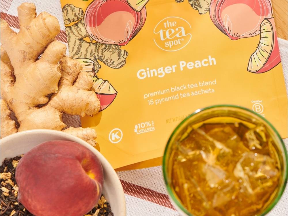 a pouch of ginger peach sachets sits on a cloth surface next to a glass of iced tea, ginger root and a bowl with loose leaf tea and peach inside