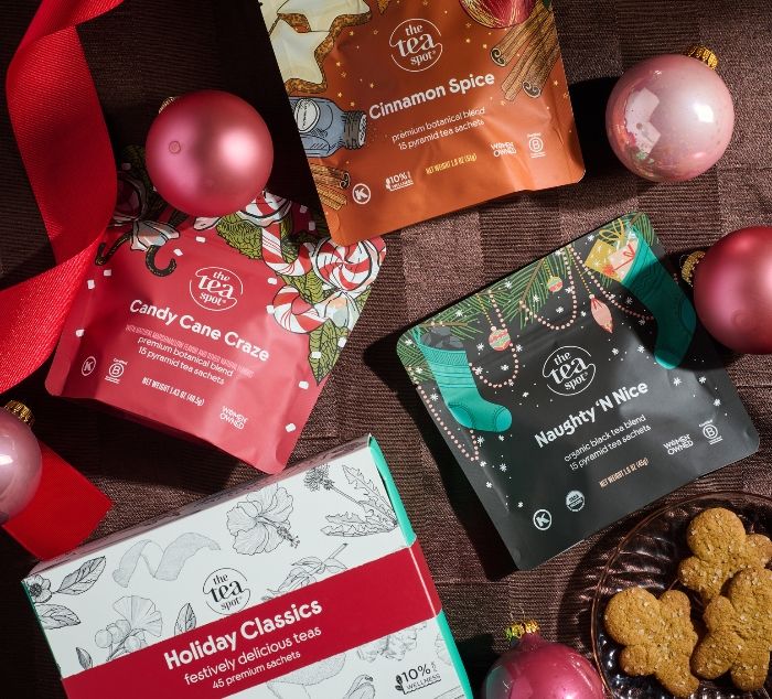 Three bags of The Tea Company holiday tea mixes arranged on a dark wood surface with pink ornaments, a red ribbon, and cookies on a plate. Visible products include Cinnamon Spice, Candy Cane Craze, Naughty 'N Nice, and the Holiday Classics box. 