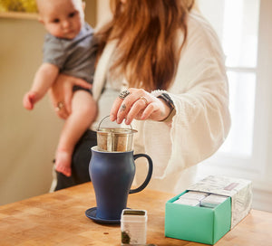 a woman is holding a baby and reaches out to take a tea infuser out of her cup on a table 