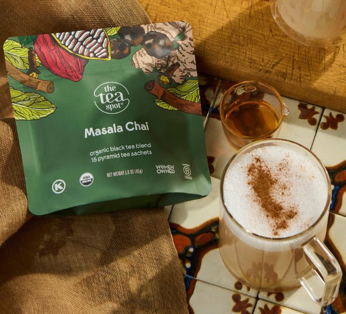 A package of The Tea Spot Masala Chai tea sachets displayed next to a frothy chai latte with cinnamon dusting on a patterned tile surface.