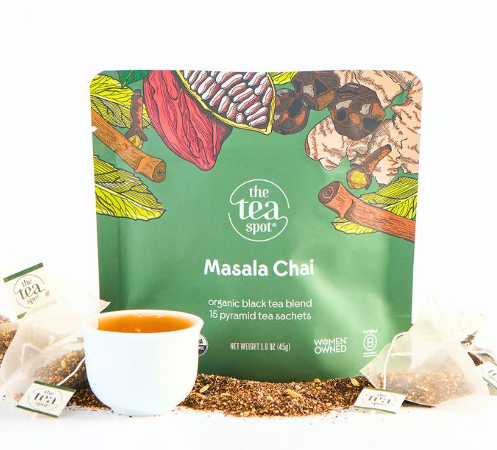A stand-up pouch of The Tea Spot Masala Chai tea blend with illustrated spices and herbs on a green background, shown with a white cup of tea and several pyramid tea sachets on a white surface.