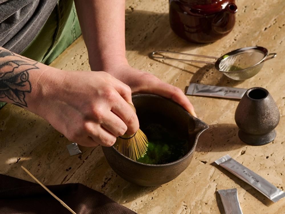 a person whisking matcha in a matcha bowl next to a matcha sifter and whisk holder