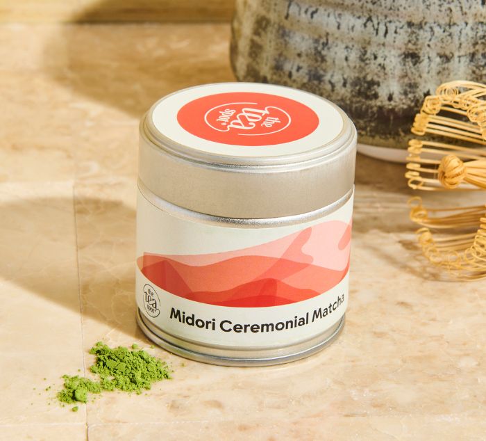 a tin of Midori Matcha sits next to a matcha whisk, bowl and matcha powder on a countertop