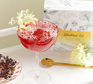 a red tea mocktail with a flower on it sits in front of a box of tea reading mocktail kit