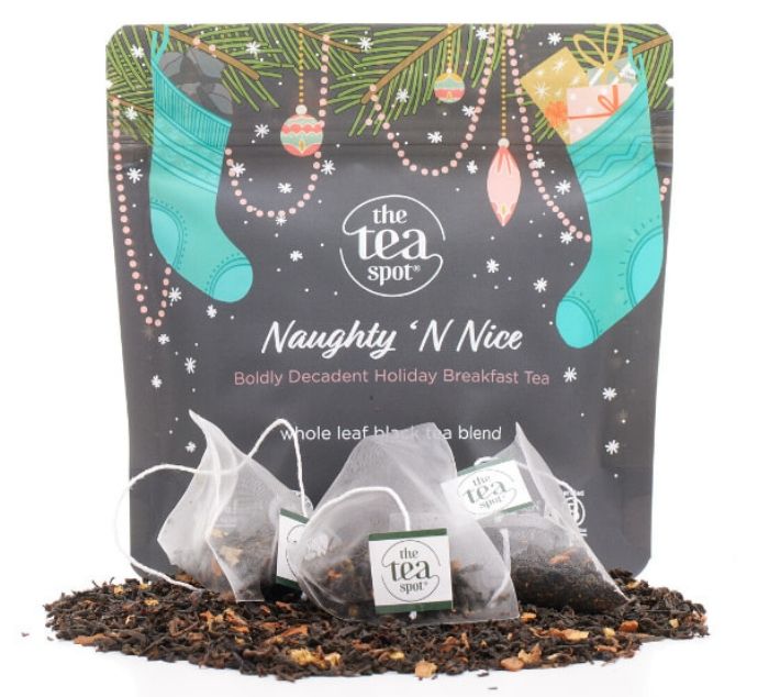The Tea Spot 'Naughty Nice' holiday breakfast tea packaging, featuring a dark gray festive pouch with teal stockings, pine branches, and ornaments, with three tea bags and loose tea leaves displayed in front on a white background.