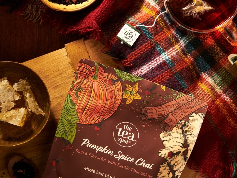 A tea bag package labeled 'Pumpkin Spice Chai' by The Tea Spot lies on a wooden table beside a glass of hot tea, a plaid blanket, and small food props in a warm autumn setting.