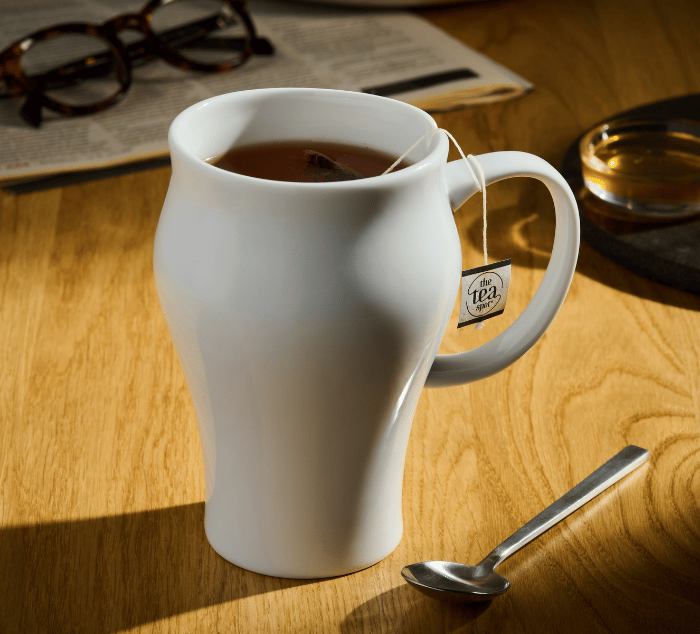 A white ceramic tea mug filled with tea and a tea bag, set on a wooden table with a spoon, glasses, and a newspaper in the background.