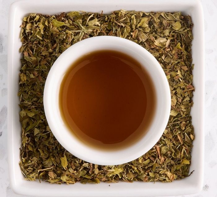 A top-down view of a white ceramic cup filled with spearmint tea, centered on a bed of dried spearmint tea leaves inside a white square dish.