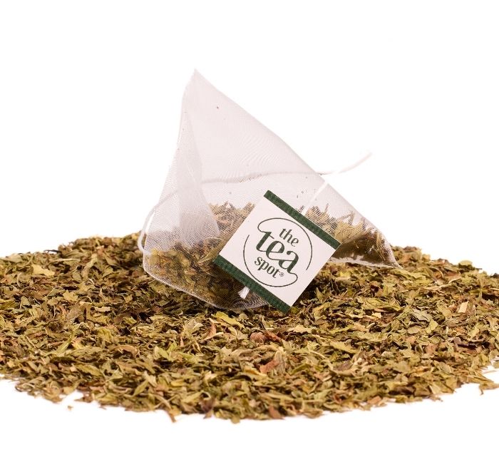 A triangular tea bag filled with loose-leaf spearmint herbal tea lies on its side on a pile of the same dried tea leaves, against a plain white background. The tag reads 'the tea spot'.