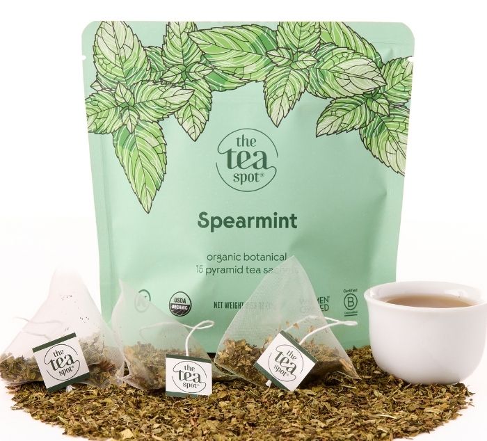 A stand-up pouch of The Tea Spot Spearmint tea bags, shown with several loose pyramid tea bags, loose spearmint leaves, and a white cup of tea on a white background.