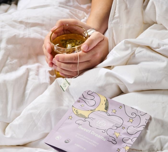 a person holds a cup of sleepy tea in bed with a package labeled sweet dreams next to them