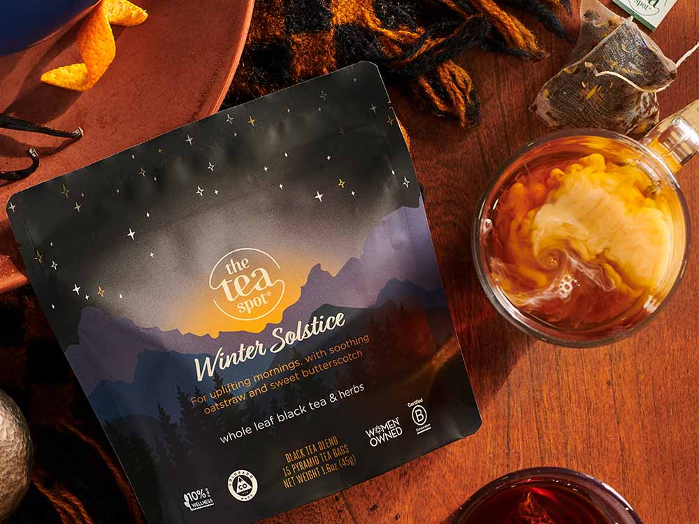 Flat lay of The Tea Spot's Winter Solstice tea packaging, featuring a serene mountain landscape design under a starry sky. Surrounding the tea bag are cozy seasonal details, including a steaming cup of tea with cream swirling in it, loose tea bags, an orange slice, and a warm knitted scarf, evoking a comforting winter atmosphere.