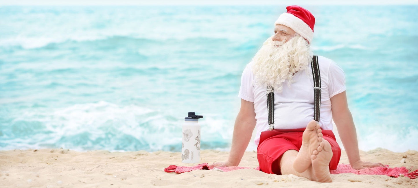 A man dressed as Santa Claus sits on a sandy beach with the ocean in the background. He is wearing a white t-shirt, red shorts, black suspenders, and a Santa hat, with bare feet visible.