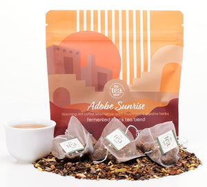 An orange bag reading adobe sunrise, coffee alternative tea with mushrooms and digestive herbs sits behind a pile of loose leaf tea and tea sachets