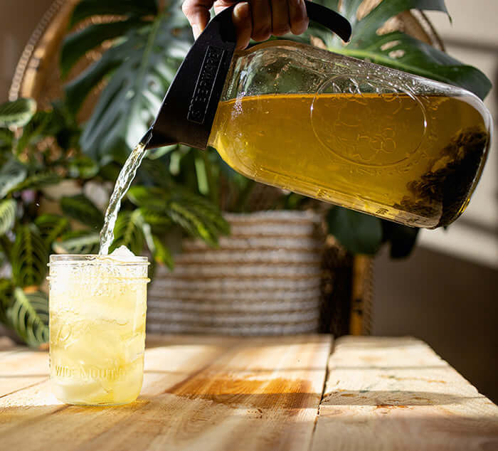 Pouring cold brew tea from a glass pitcher into a mason jar on a wooden table, surrounded by lush greenery.