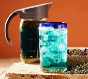 a cup with iced tea in it sits in front of a mason jar with blue tea brewed in it on a rock