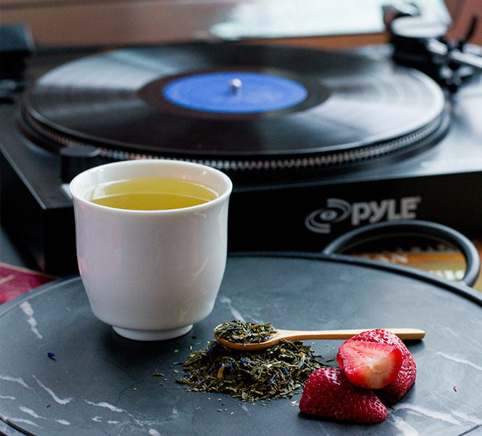 
                  
                    a cup of green tea sits in front of a record player with loose leaf tea and strawberries around it
                  
                