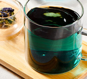 a cup with a butterfly pea flower blend brewed in it sits on a wooden board with loose leaf tea next to it