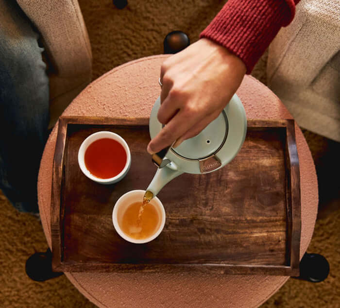 Hand pouring tea into ceramic teacups on wooden tray, showcasing loose leaf tea service for two.