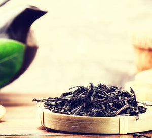 Black sundried tea leaves are piled on a coaster in front of a black teapot